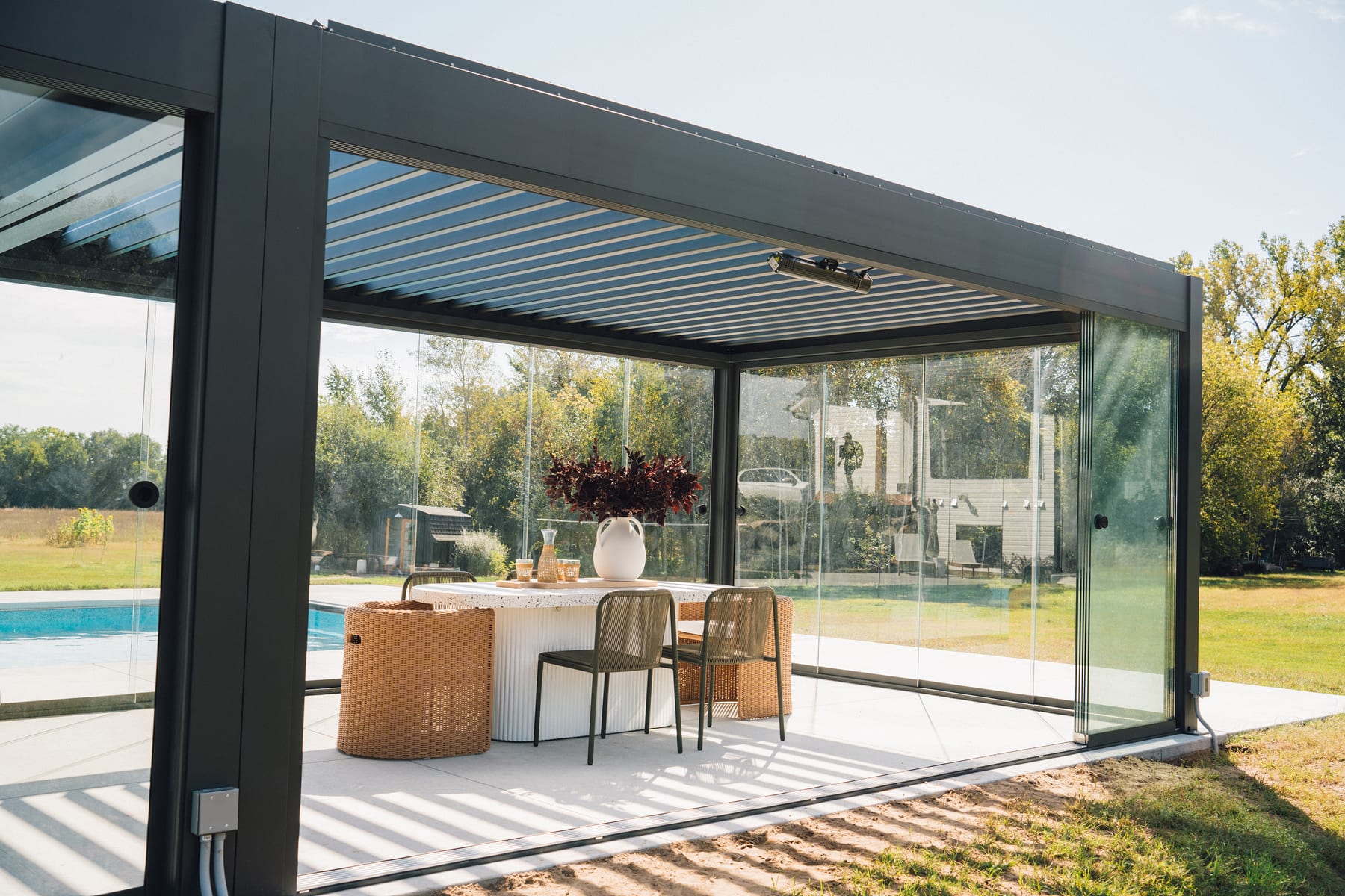 Anthracite grey louvered-roof pergola with sliding glass walls enclosing a dining set beside a residential pool on a sunny day.