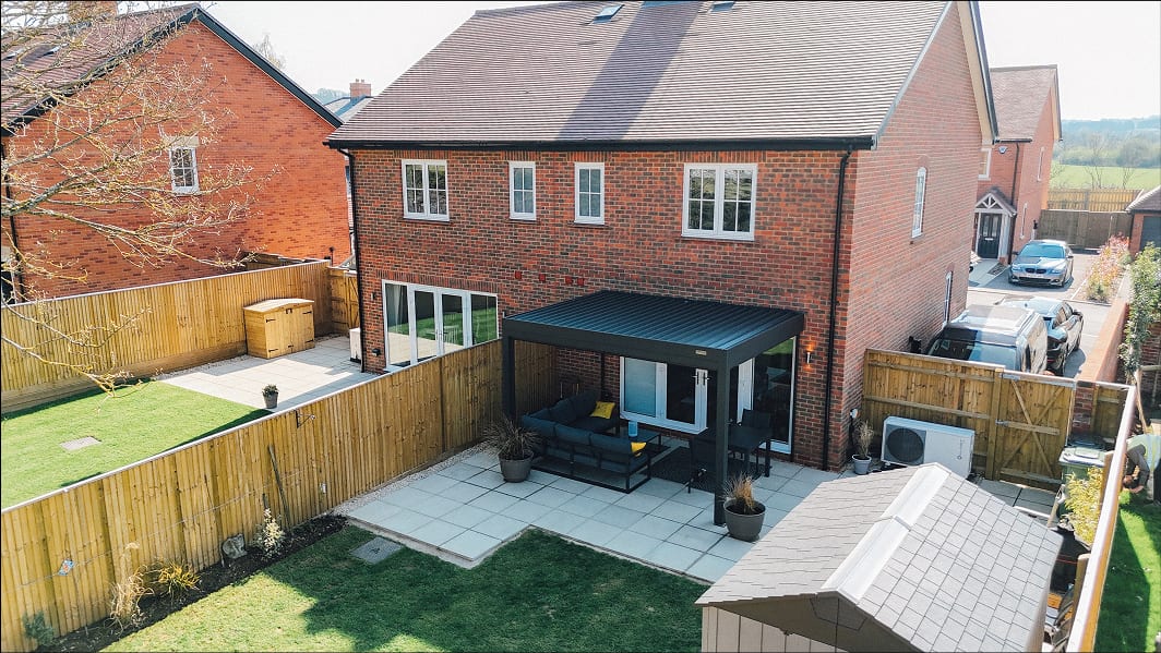 Anthracite grey louvered pergola attached to a red-brick house rear, with patio sofa set beneath, viewed from above.