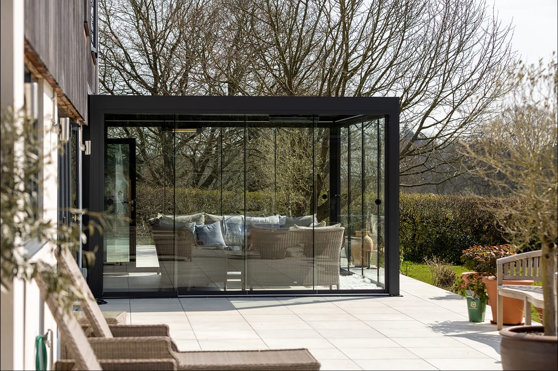 Anthracite grey aluminium pergola with sliding glass walls enclosing wicker lounge furniture on a light stone patio, garden and bare trees in background.
