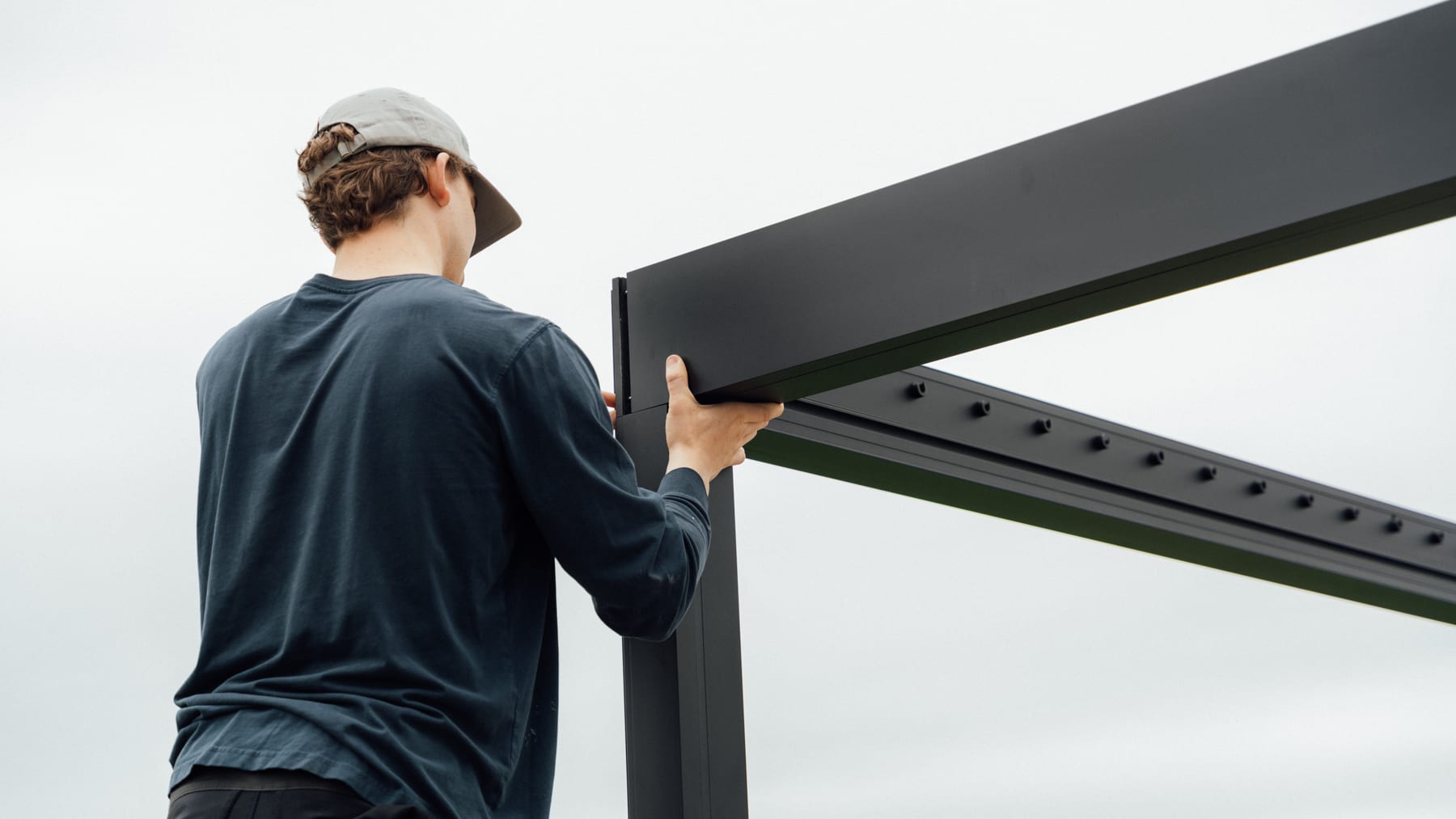 Two people assembling an aluminium louvered pergola frame in a British back garden, one person on a ladder fitting a motorised slat
