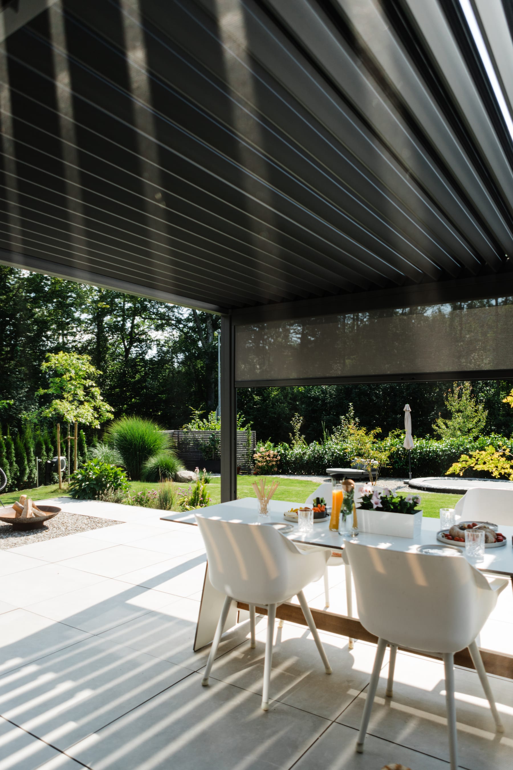 Anthracite-grey bioclimatic pergola with louvered roof and roller blind over a set white outdoor dining table on a tiled garden patio.