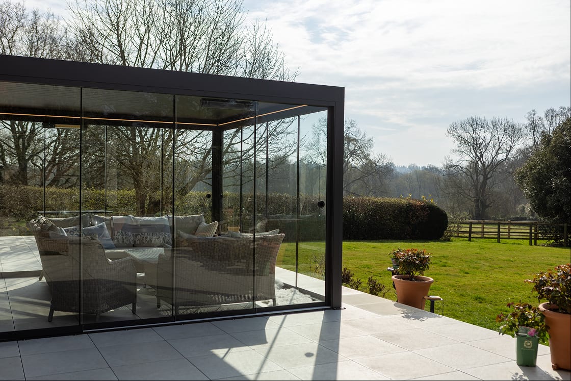 Anthracite-grey aluminium pergola with sliding glass walls and rattan sofas on a paved patio, set against a green rural garden landscape.
