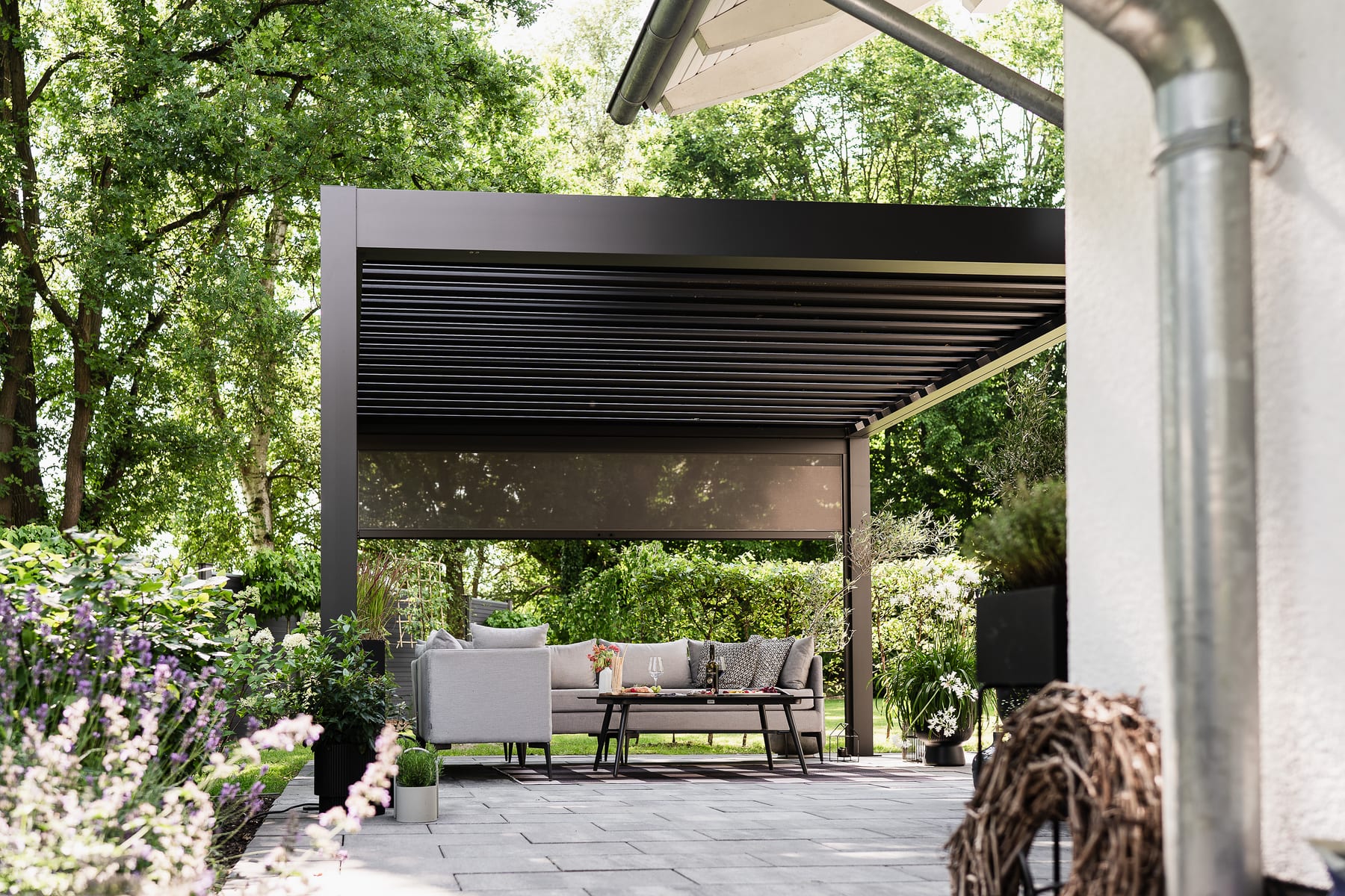 Anthracite-grey aluminium louvered pergola with roller blind screen and grey outdoor sofa set on a stone patio in a green residential garden, midday light.
