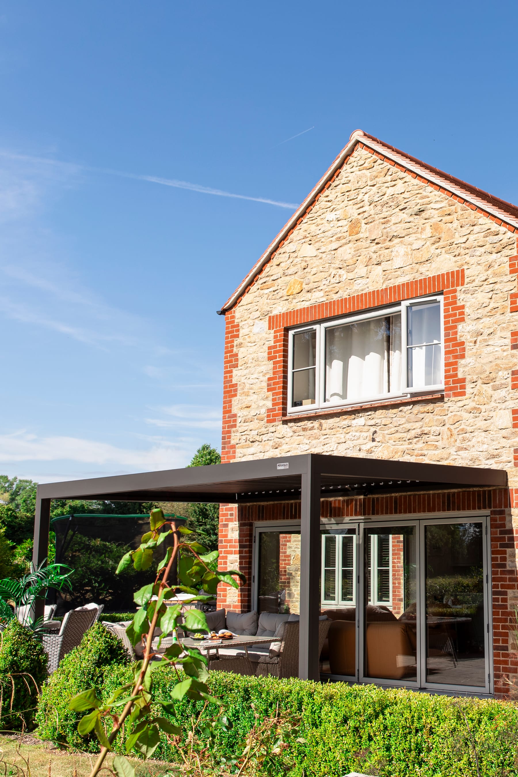 UK garden with an L-shaped patio and a PERGOLUX Custom Design aluminium pergola fitted around a mature tree, showing a non-standard footprint