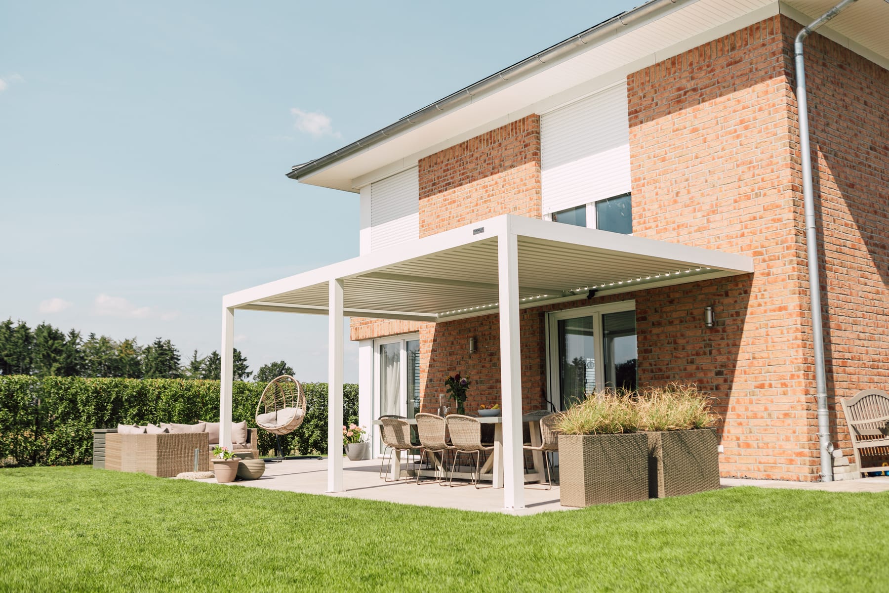 White bioclimatic louvered pergola on a sunny residential patio with rattan dining furniture, hanging egg chair, and ornamental planters.